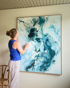 Danish artist contemplating her large original abstract painting on white wall – expressive square canvas artwork in teal, deep blue and white with fluid brushwork and paint splatter, in natural oak float frame, with Wishbone chair in foreground