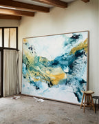 Large abstract painting on canvas in teal, blue and gold tones with oak frame, leaning against a white wall in a rustic artist's studio with exposed wooden ceiling beams and concrete floor