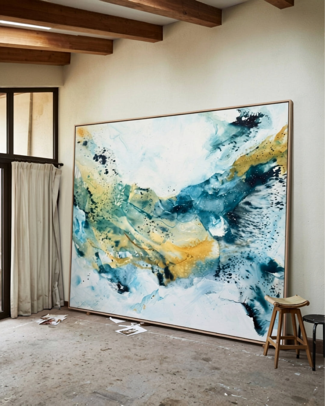 Large abstract painting on canvas in teal, blue and gold tones with oak frame, leaning against a white wall in a rustic artist's studio with exposed wooden ceiling beams and concrete floor