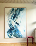 Large original abstract painting in vertical format hanging on white wall in Scandinavian living room – expressive canvas artwork in teal, cobalt blue and white with fluid brushwork and fine paint details, in natural oak float frame, with red stool and parquet floor