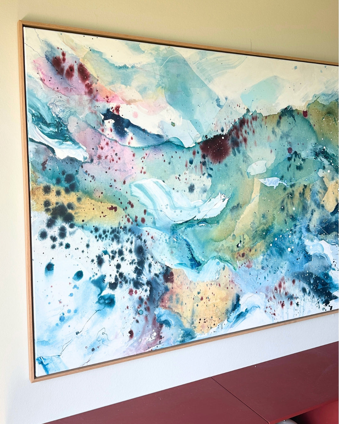 Large original abstract painting in horizontal format hanging on warm beige wall above red Montana shelving unit – expressive canvas artwork in teal, sky blue, blush pink, gold and white with fluid brushwork, in natural oak float frame