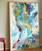 Soft #12 – original abstract canvas painting, 100x150 cm, in natural oak frame. Flowing layers of teal, gold, blue, rose and burgundy by Danish artist Mette Lindberg, shown in Scandinavian home interior