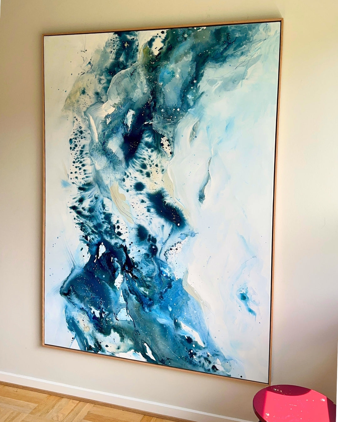 Large original abstract painting in vertical format hanging on white wall in Scandinavian room – expressive canvas artwork in teal, cobalt blue, navy and white with fluid dynamic brushwork, in natural oak float frame, with pink stool and parquet floor
