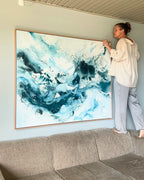 Danish artist hanging large original abstract painting above sofa on sage green wall – expressive square canvas artwork in teal, deep blue and white with fluid brushwork and paint splatter, in natural oak float frame, shown in Scandinavian living room