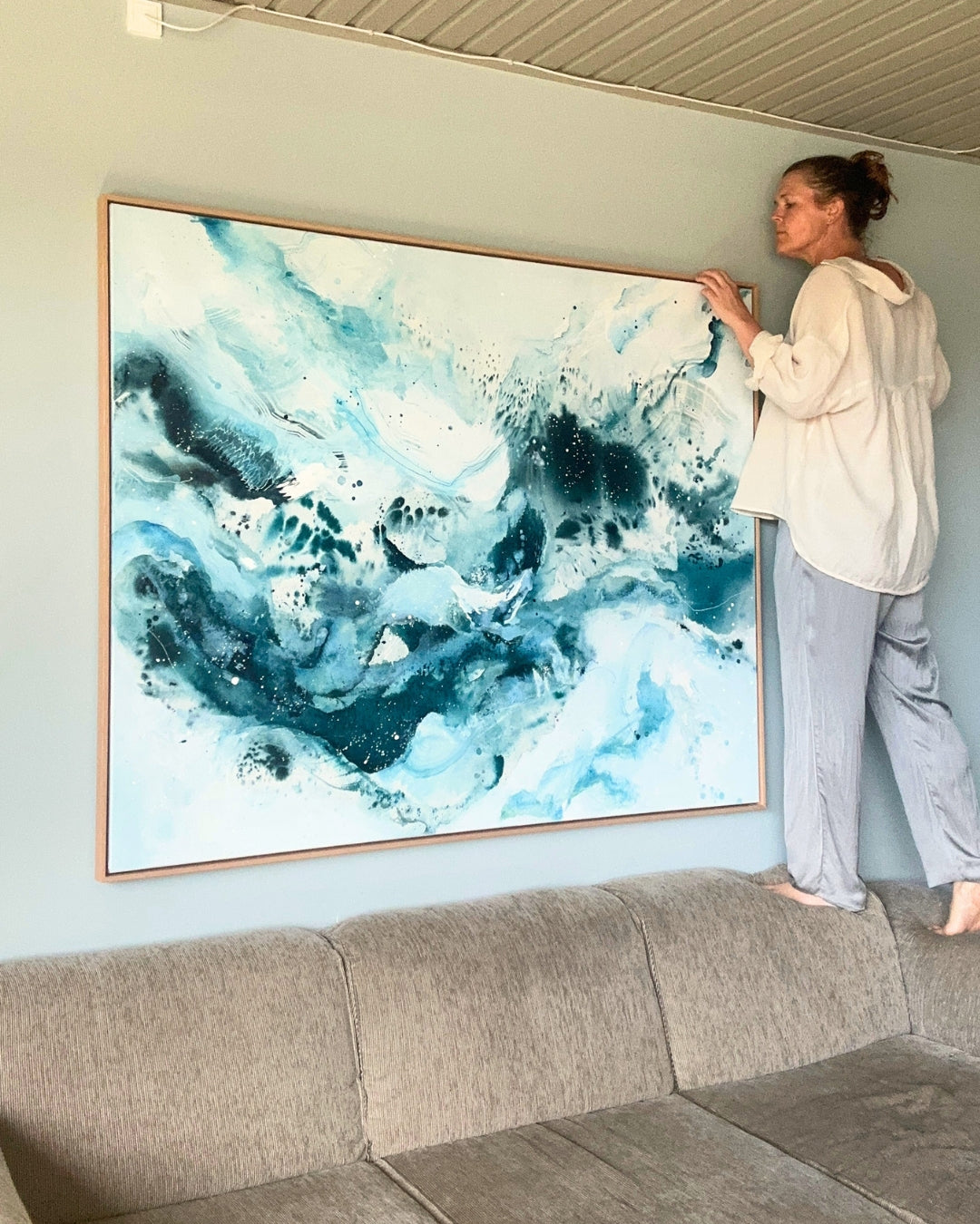 Danish artist hanging large original abstract painting above sofa on sage green wall – expressive square canvas artwork in teal, deep blue and white with fluid brushwork and paint splatter, in natural oak float frame, shown in Scandinavian living room