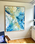 Original abstract canvas painting in deep teal, cobalt blue and warm yellow tones, in natural oak float frame – dynamic paint splatter and fluid brushwork by Danish contemporary artist Mette Lindberg, shown in Scandinavian interior