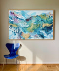 Soft #12 – original abstract canvas painting, 100x150 cm, in natural oak frame. Expressive fluid layers in teal, blue, gold and rose tones by Danish artist Mette Lindberg, shown in Scandinavian interior.