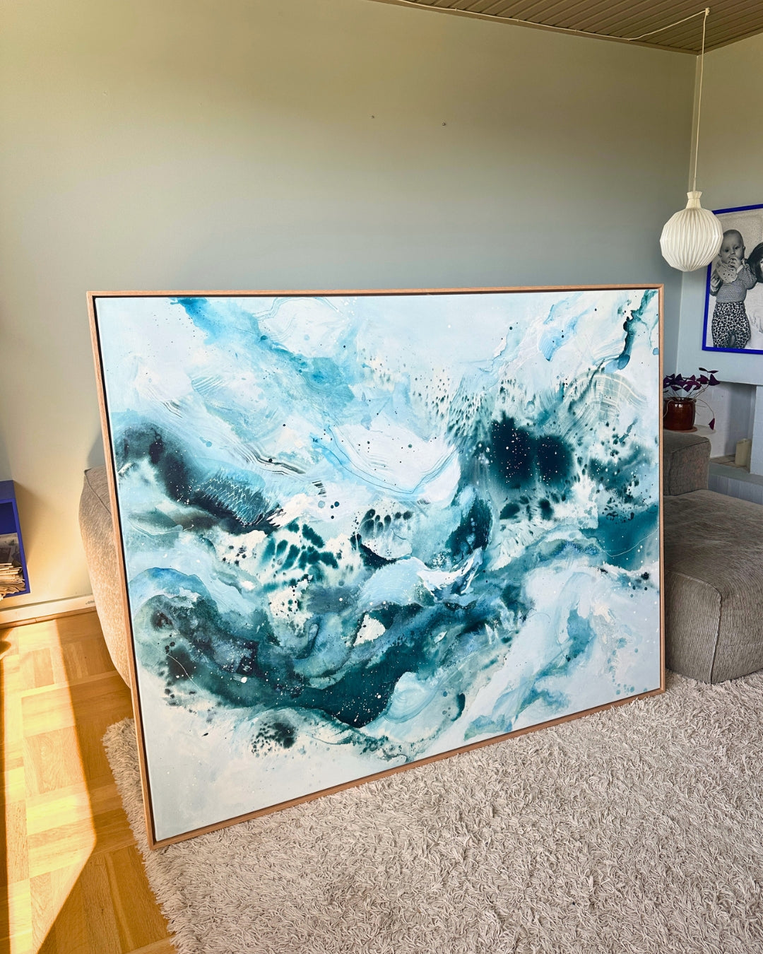 Large original abstract painting leaning against wall in Scandinavian living room – expressive canvas artwork in teal, deep blue, white and soft grey tones with fluid brushwork and paint splatter, in natural oak float frame