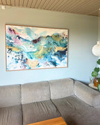 Large original abstract painting in horizontal format hanging above corner sofa on sage green wall – expressive canvas artwork in teal, sky blue, blush pink, gold and white with fluid brushwork, in natural oak float frame, shown in Scandinavian living room with pendant lamp and monstera plant.