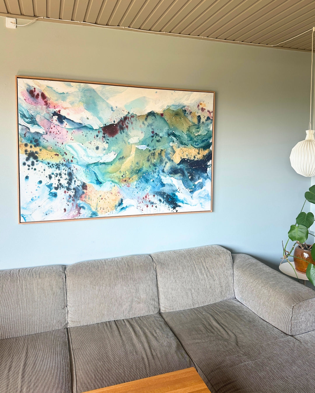 Large original abstract painting in horizontal format hanging above corner sofa on sage green wall – expressive canvas artwork in teal, sky blue, blush pink, gold and white with fluid brushwork, in natural oak float frame, shown in Scandinavian living room with pendant lamp and monstera plant.