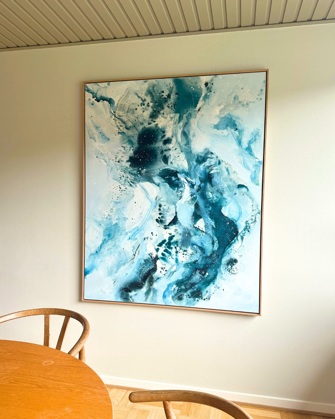 Large original abstract painting hanging on white wall above oak dining table and Wishbone chair – expressive canvas artwork in teal, cobalt blue and white with fluid brushwork, in natural oak float frame, shown in Scandinavian dining room with parquet floor