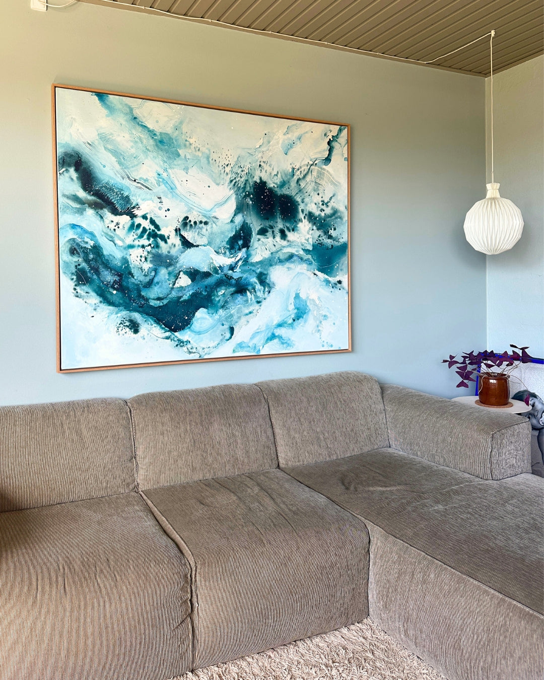 Large original abstract painting hanging above corner sofa on sage green wall – expressive canvas artwork in teal, deep blue and white with fluid brushwork and paint splatter, in natural oak float frame, shown in Scandinavian living room with pendant lamp and purple plant