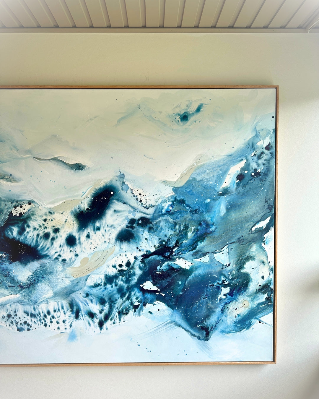 Large original abstract painting in horizontal format hanging on white wall in bright Scandinavian room – expressive canvas artwork in teal, cobalt blue and white with fluid dynamic brushwork, in natural oak float frame, photographed from below