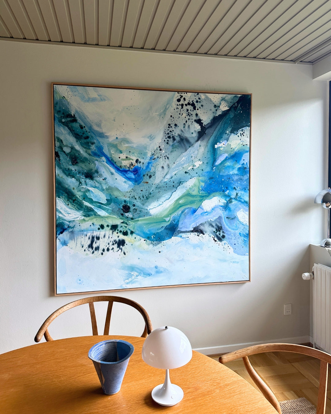 Large square original abstract painting leaning against wall on parquet floor, in natural oak float frame – expressive fluid layers in teal, cobalt blue, sage green and white with dynamic black paint splatter by Danish artist Mette Lindberg