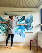 Danish artist arranging two large original abstract paintings on white wall – expressive canvas artworks in teal, cobalt blue, sage green and white with black paint splatter, in natural oak float frames, shown in Scandinavian interior with parquet floor