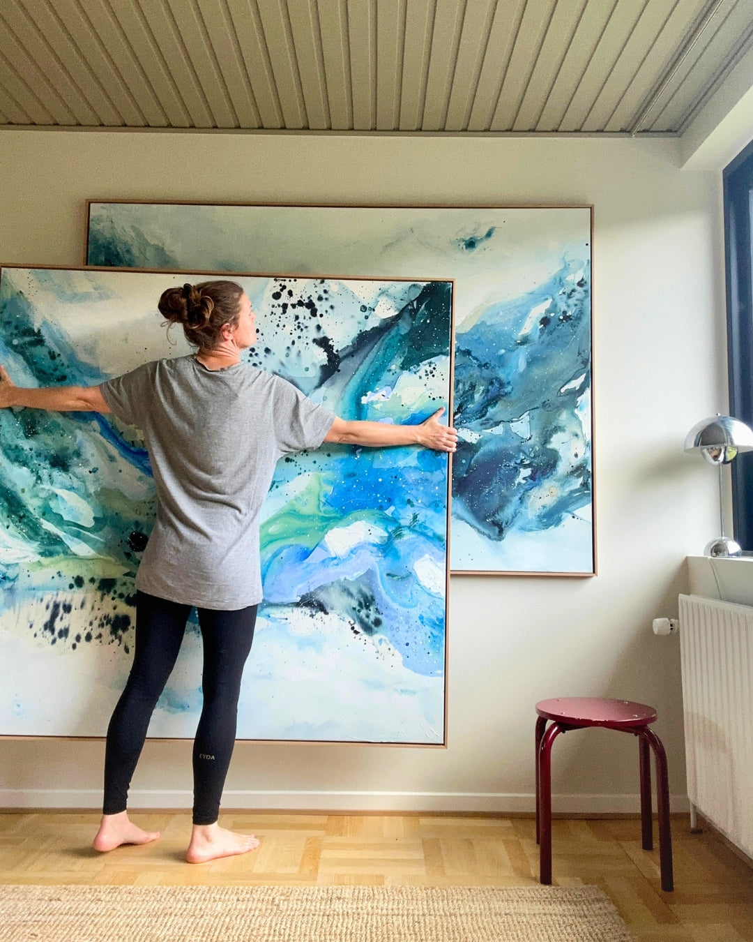 Danish artist arranging two large original abstract paintings on white wall – expressive canvas artworks in teal, cobalt blue, sage green and white with black paint splatter, in natural oak float frames, shown in Scandinavian interior with parquet floor