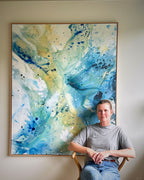 Danish artist Mette Lindberg seated in front of her large original abstract painting – expressive canvas artwork in teal, cobalt blue and warm yellow tones with fluid brushwork and paint splatter, in natural oak frame