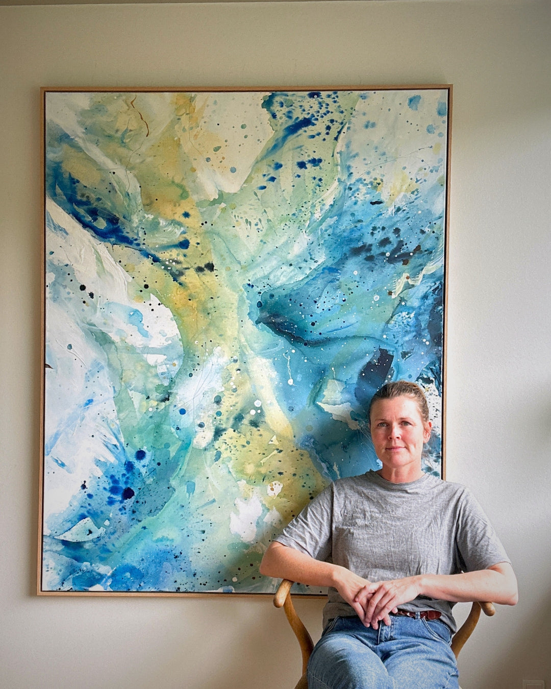 Danish artist Mette Lindberg seated in front of her large original abstract painting – expressive canvas artwork in teal, cobalt blue and warm yellow tones with fluid brushwork and paint splatter, in natural oak frame