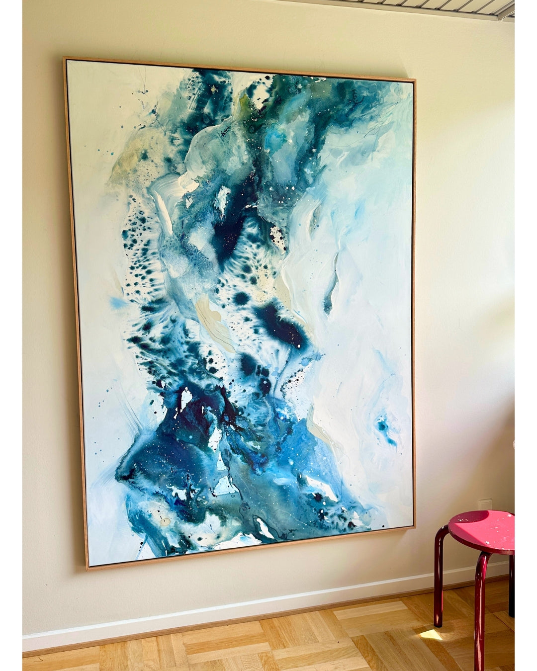 Large original abstract painting in vertical format hanging on white wall in Scandinavian room – expressive canvas artwork in teal, cobalt blue, navy and white with fluid dynamic brushwork, in natural oak float frame, with red stool and herringbone parquet floor