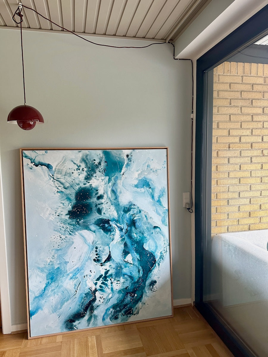 Original abstract canvas painting, 120x150 cm, in natural oak frame. Fluid and expressive teal, deep blue and white tones by Danish artist Mette Lindberg, shown leaning against wall in Danish home
