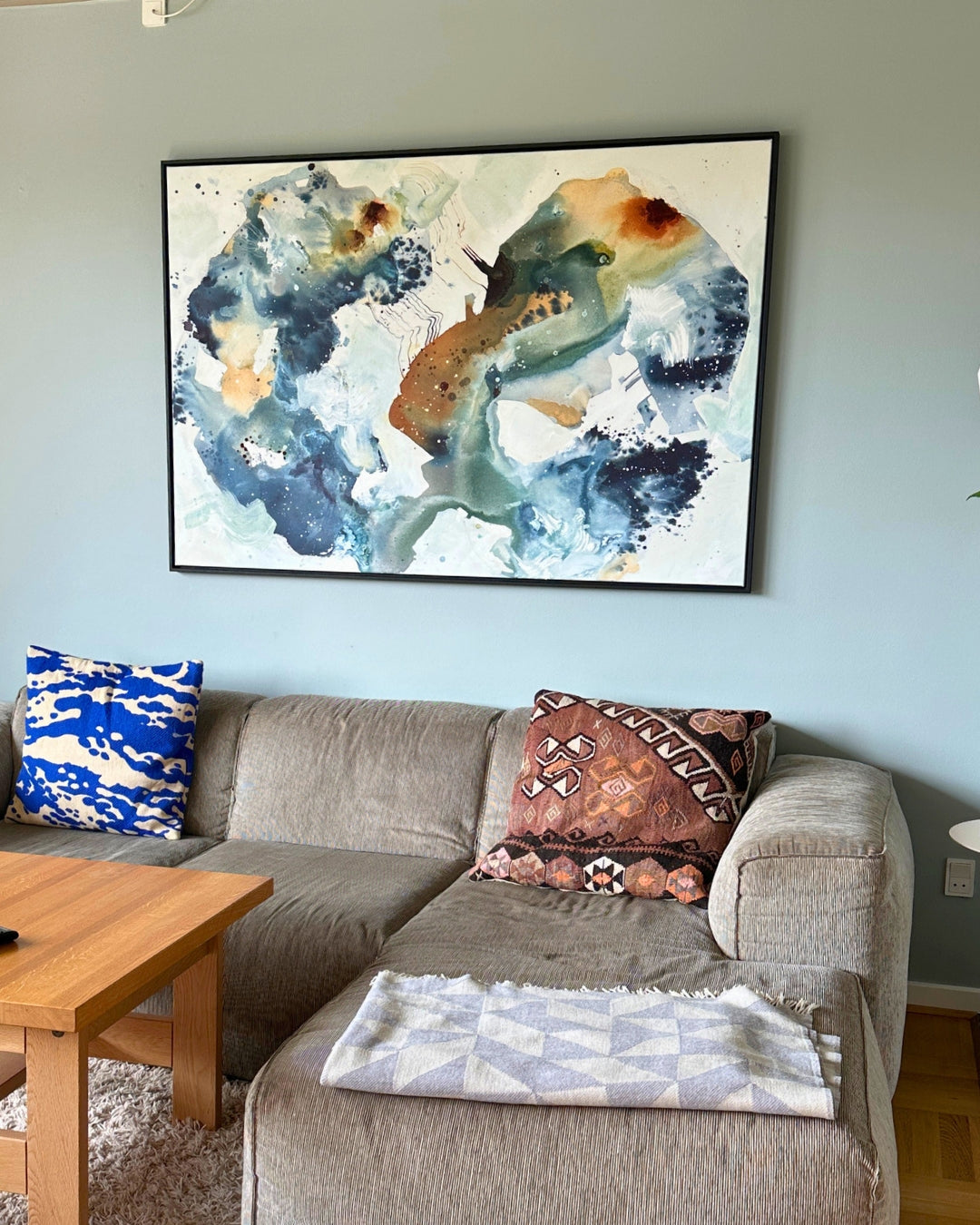 Large abstract painting in blue, green, and warm brown tones hanging above a grey corner sofa against a light blue wall. The painting is framed in black and measures 100×150 cm.