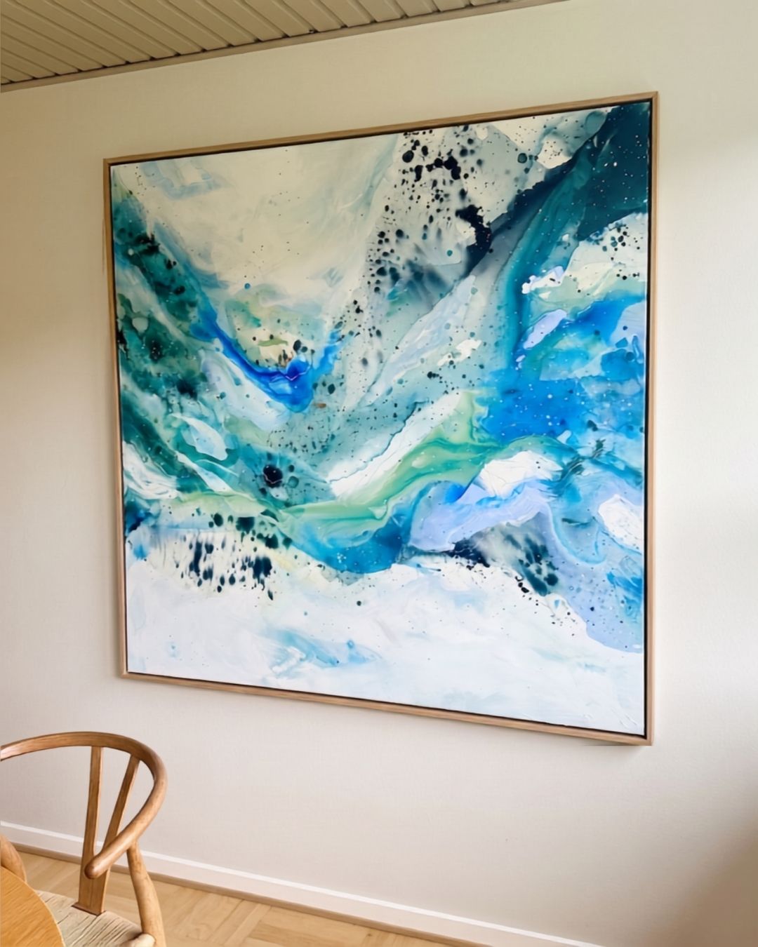 Large abstract painting on canvas in flowing shades of teal, blue, green and white with dark ink splatter details, framed in natural oak, hung on a white wall above a light wood chair