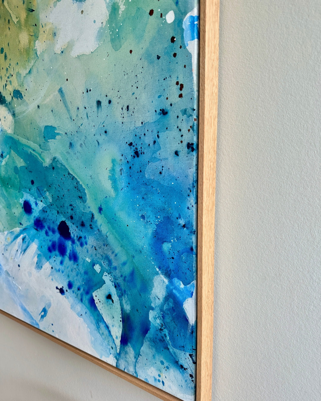 Close-up detail of an abstract painting in shades of blue, turquoise, and green with dark ink splatter, framed in natural oak. The textured brushwork and fluid layers give the work an organic, water-like quality