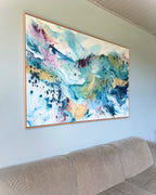Large original abstract painting in horizontal format hanging above sofa on sage green wall – expressive canvas artwork in teal, sky blue, blush pink, gold and white with fluid brushwork, in natural oak float frame, shown in Scandinavian living room.