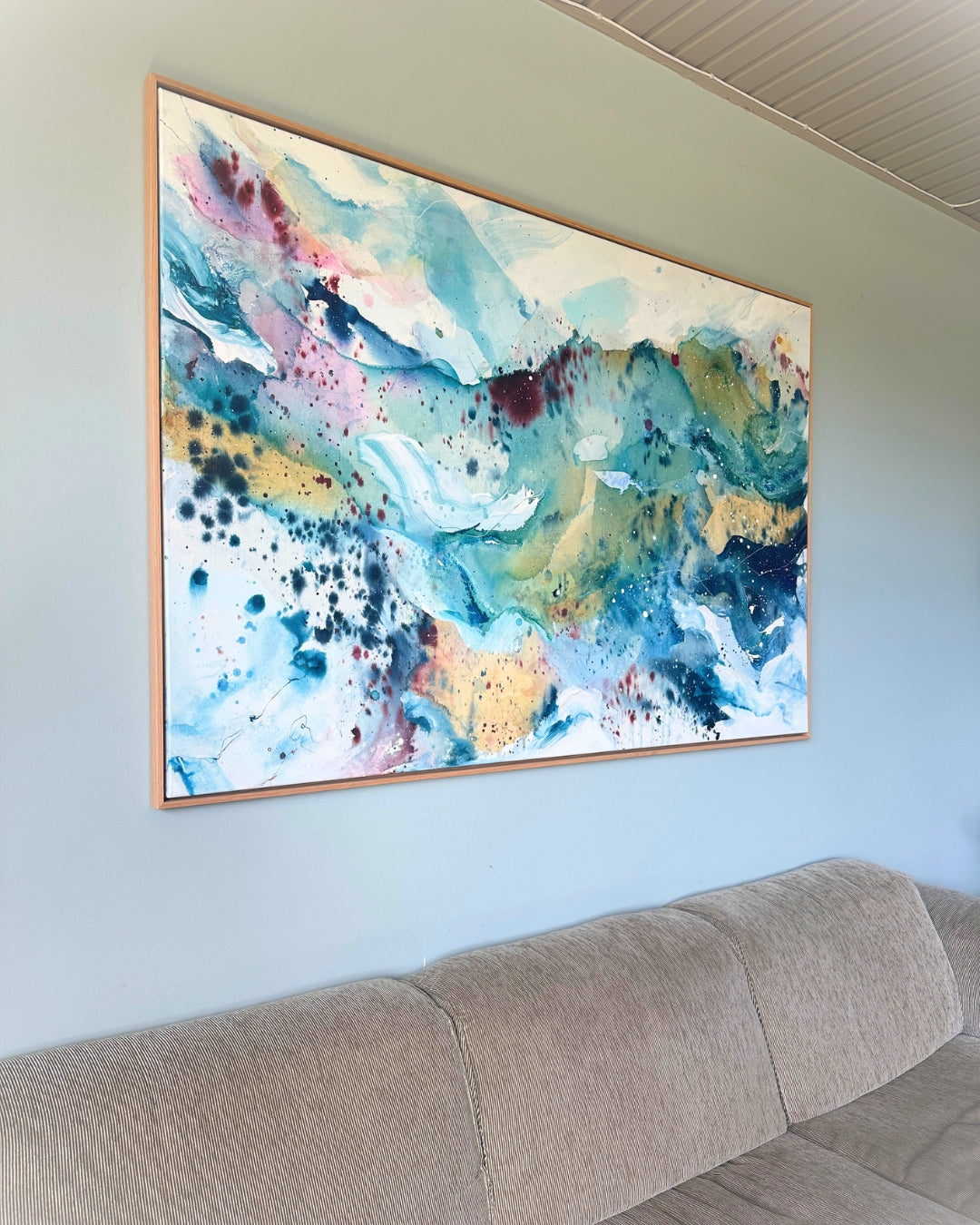 Large original abstract painting in horizontal format hanging above sofa on sage green wall – expressive canvas artwork in teal, sky blue, blush pink, gold and white with fluid brushwork, in natural oak float frame, shown in Scandinavian living room.