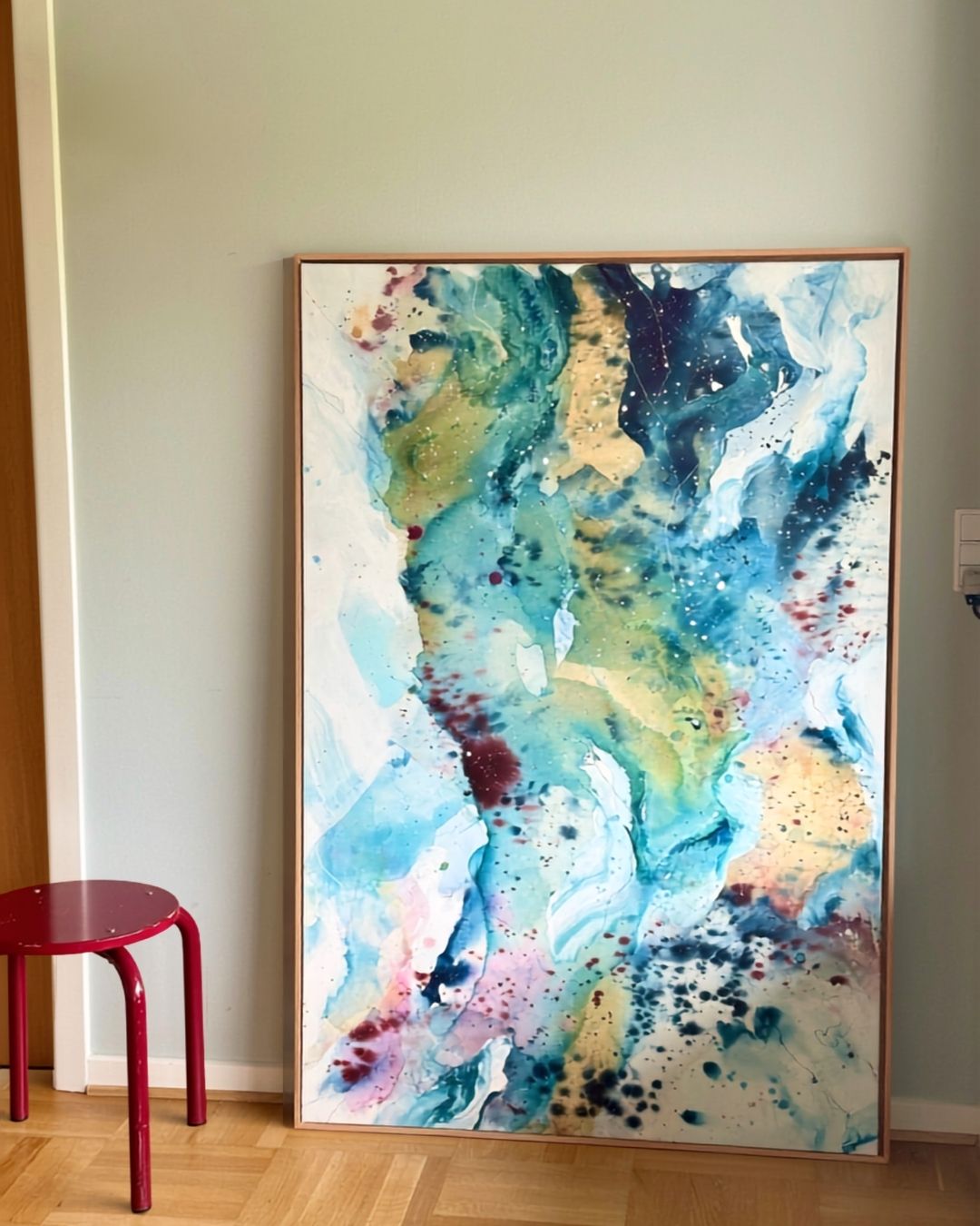 Tall abstract painting on canvas in flowing teal, aqua, gold, purple and white with scattered ink drops in burgundy and dark blue, framed in natural oak, leaning against a sage green wall on a parquet floor with a red metal stool beside it