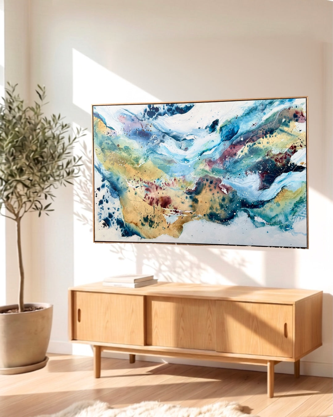 Large horizontal abstract painting with flowing blue, teal, gold and burgundy tones with ink splashes, displayed in a natural wood frame above a Scandinavian sideboard with an olive tree beside it