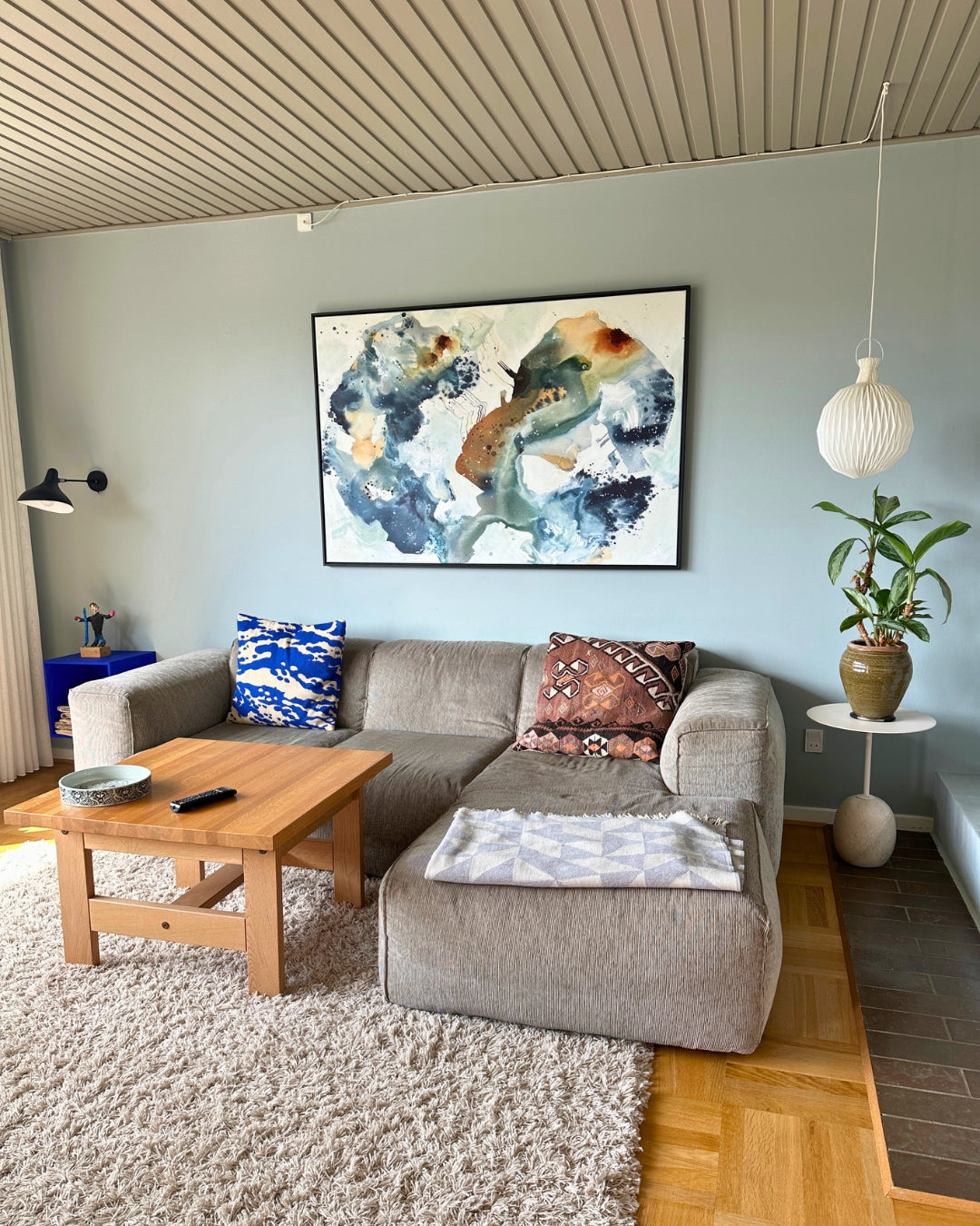 Large abstract painting in shades of blue, green, and brown hanging above a grey sofa in a bright living room with Nordic interior design. The painting is framed in black and measures 100×150 cm