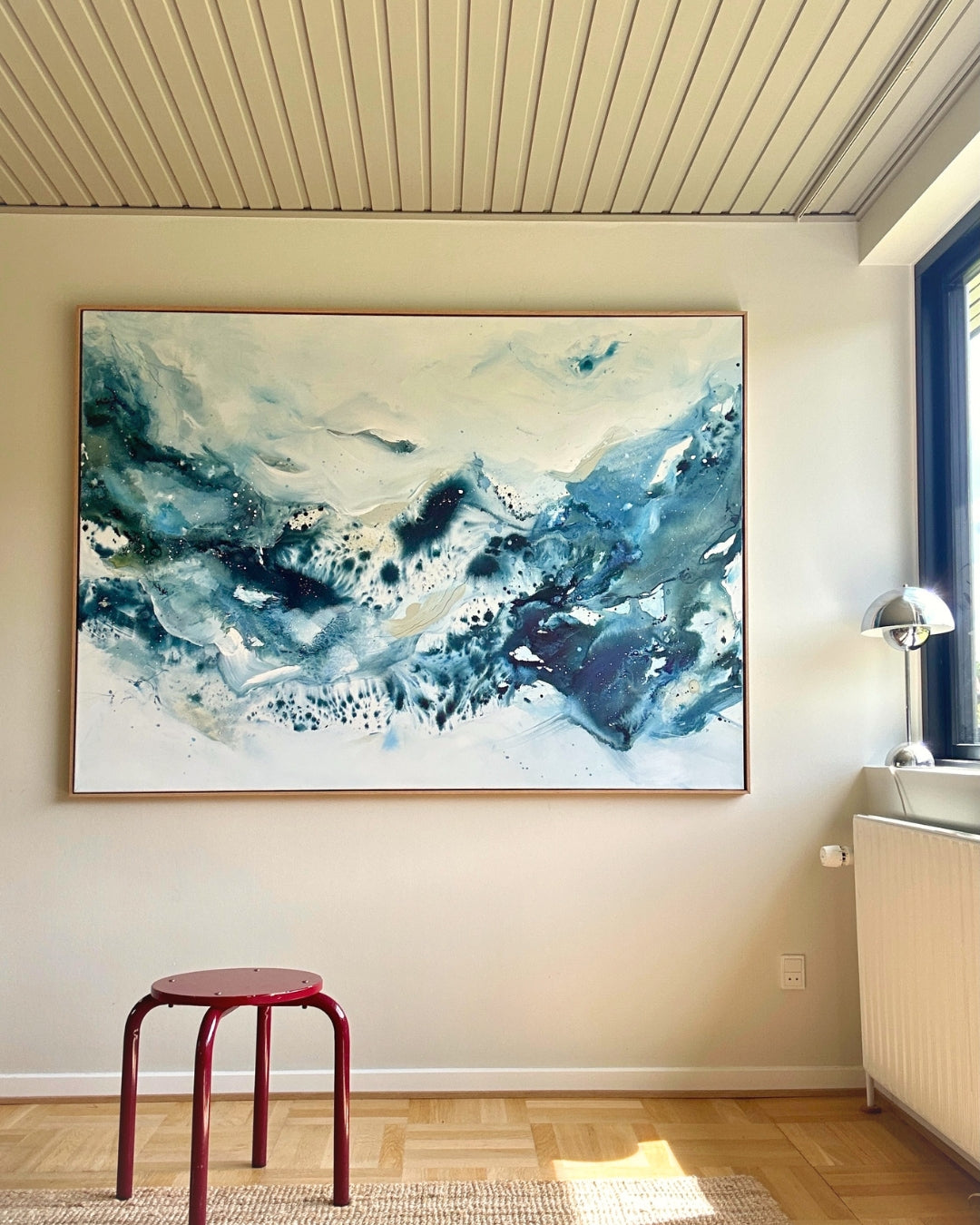 Large original abstract painting in horizontal format hanging on white wall in bright Scandinavian room – expressive canvas artwork in teal, cobalt blue and white with fluid brushwork, in natural oak float frame, with red stool, chrome floor lamp and parquet floor