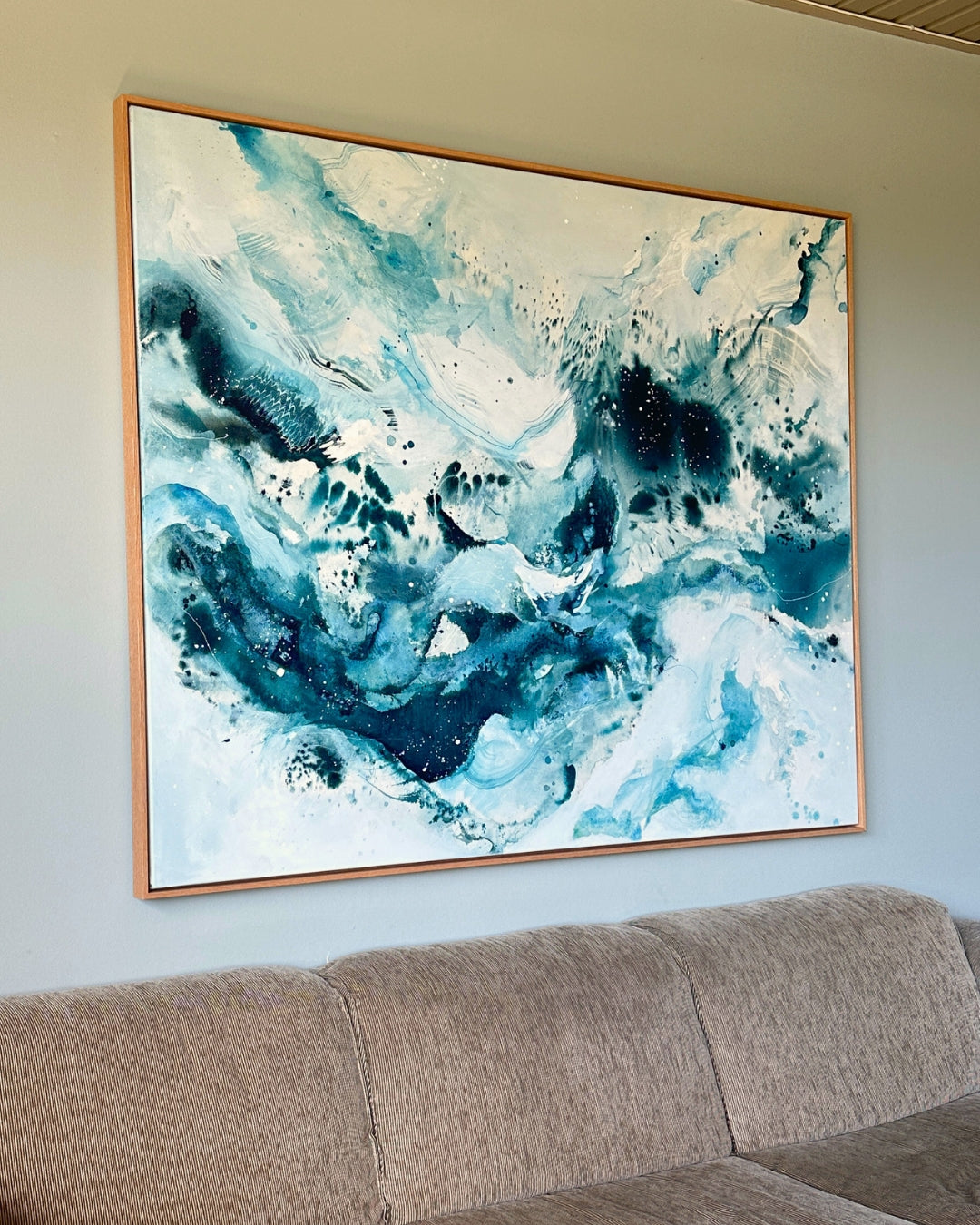 Large original abstract painting hanging above sofa on sage green wall – expressive square canvas artwork in teal, cobalt blue and white with dynamic fluid brushwork and paint splatter, in natural oak float frame, shown in Scandinavian living room