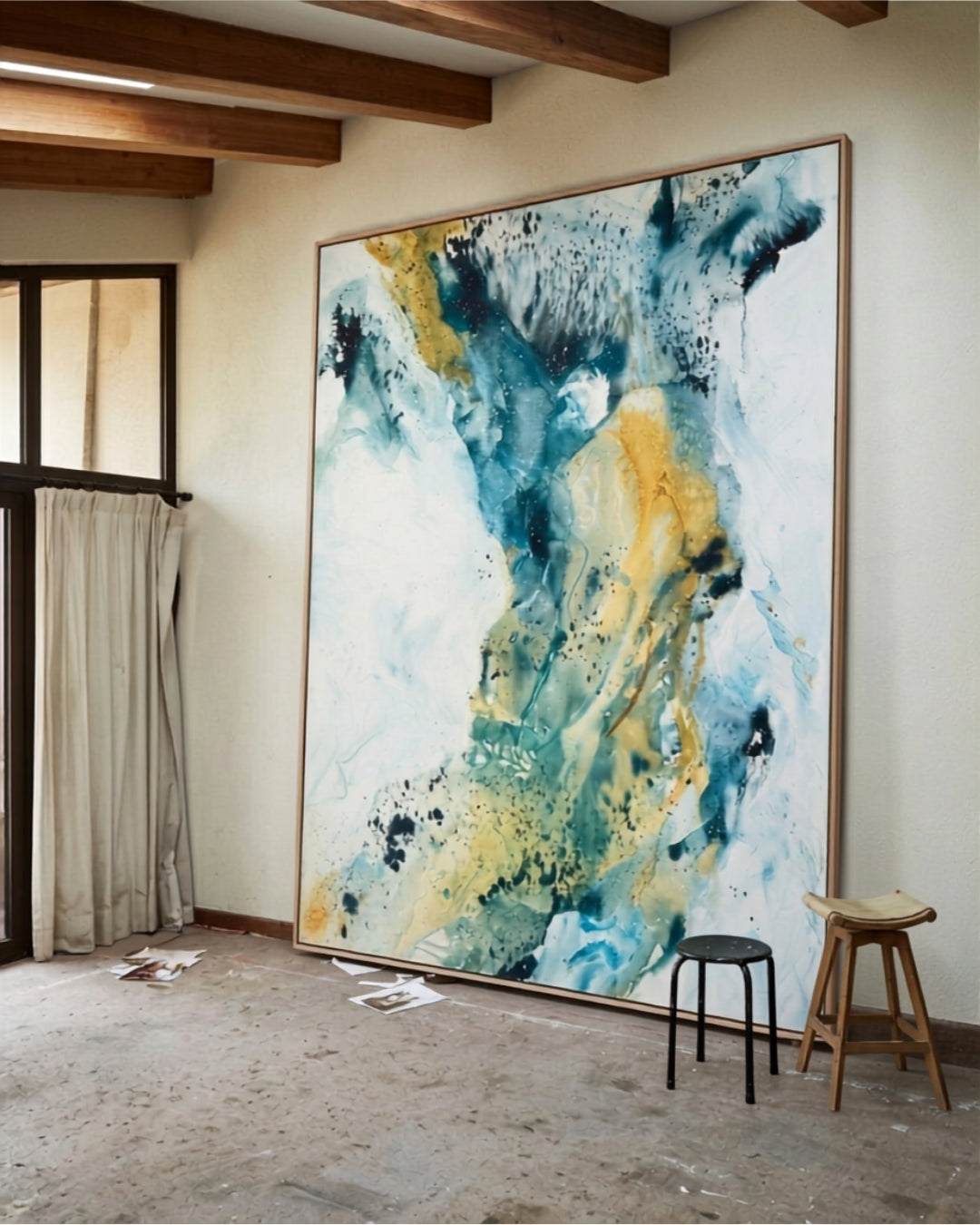 Monumental abstract painting on canvas in teal, blue and gold tones with oak frame, standing against a white wall in a rustic artist's studio with exposed wooden ceiling beams and concrete floor