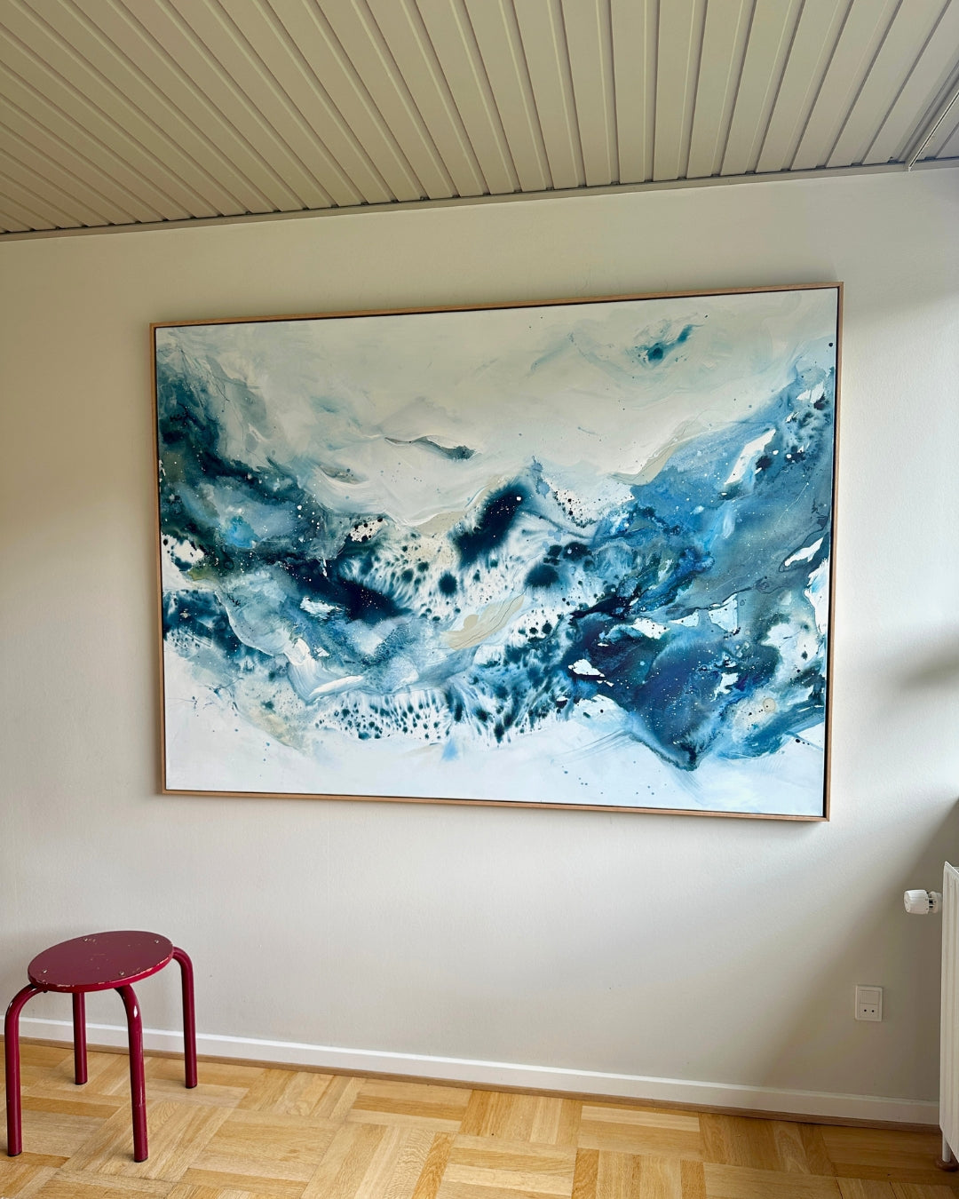 Large original abstract painting in horizontal format hanging on white wall in Scandinavian living room – expressive canvas artwork in teal, cobalt blue and white with fluid brushwork and paint splatter, in natural oak float frame, with red stool and parquet floor