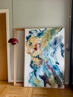 Soft #13 – original abstract canvas painting, 100x150 cm, in natural oak frame. Expressive fluid layers in teal, gold, blue and burgundy tones by Danish artist Mette Lindberg, shown leaning against wall in Nordic home