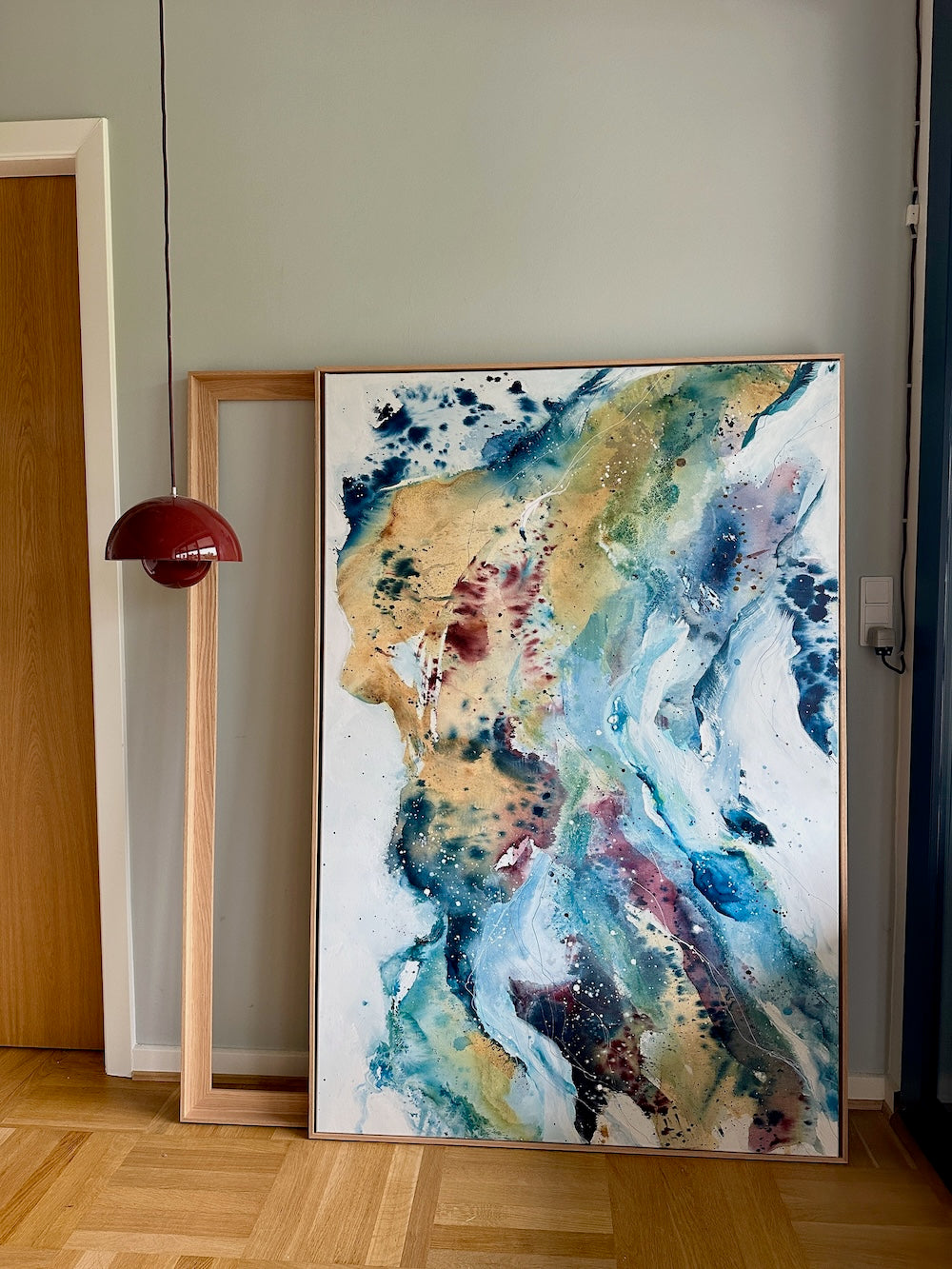 Soft #13 – original abstract canvas painting, 100x150 cm, in natural oak frame. Expressive fluid layers in teal, gold, blue and burgundy tones by Danish artist Mette Lindberg, shown leaning against wall in Nordic home