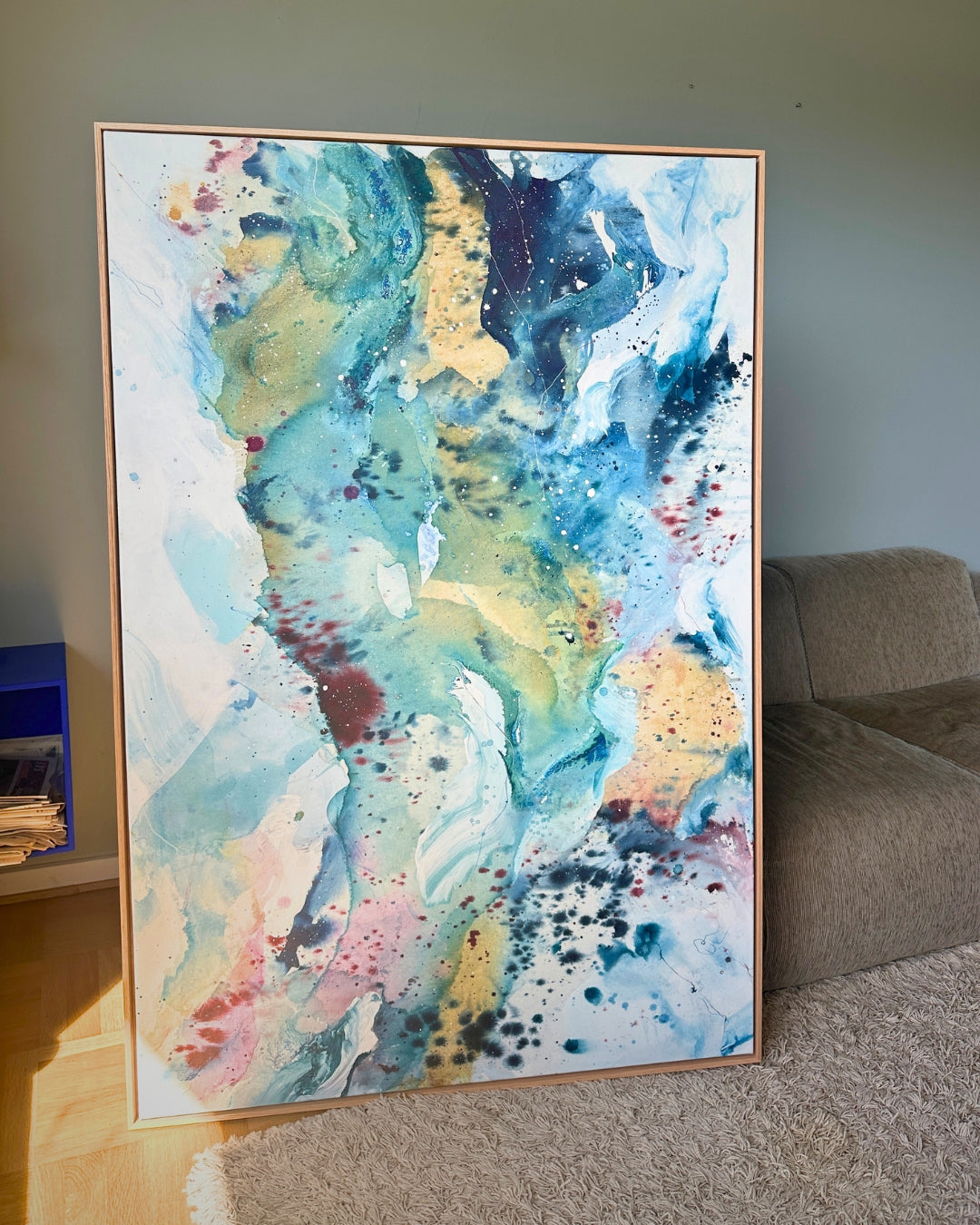 Large original abstract painting in vertical format leaning against sage green wall in Scandinavian living room – expressive canvas artwork in teal, sky blue, gold, blush pink and white with fluid brushwork, in natural oak float frame, with sofa and parquet floor