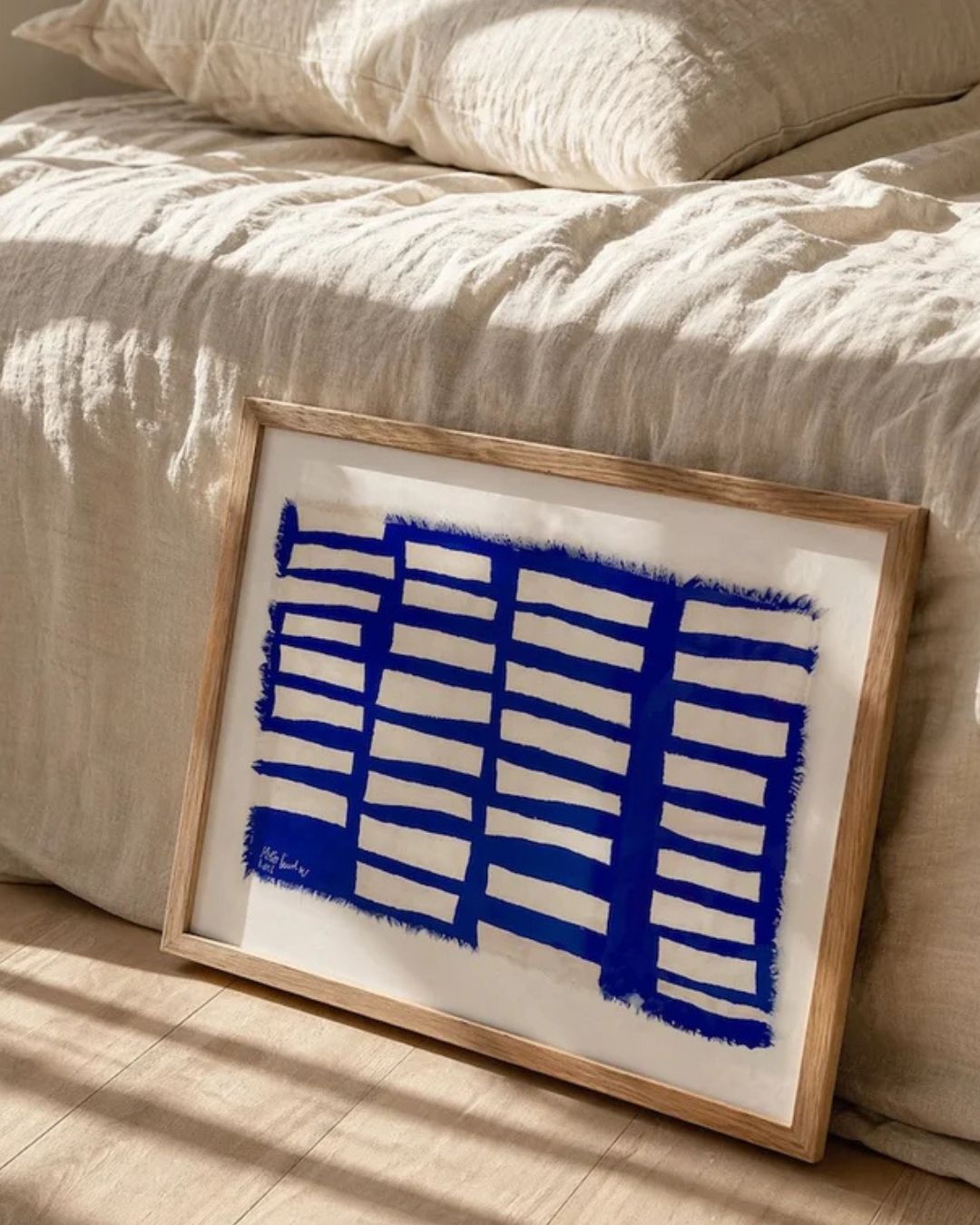 Abstract painting with bold ultramarine blue grid pattern on a white background, framed in natural oak, leaning against a bed with linen bedding in warm neutral tones